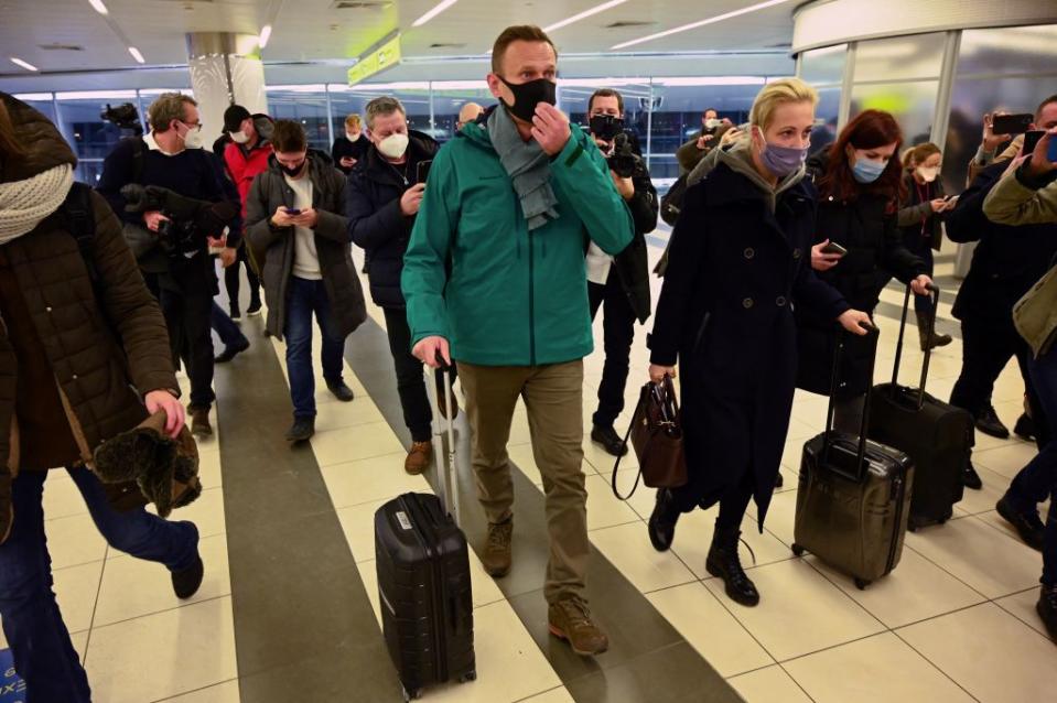 alexei navalny and yulia navalnaya walk on a tiled floor while rolling black suitcases and wearing face masks, coats and pants, a group of people walks alongside and behind them