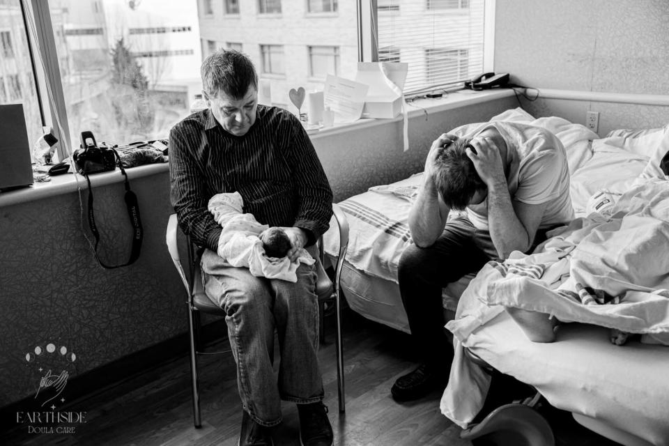 Two adults and a newborn in a hospital room, one seated holding baby, the other with head in hands