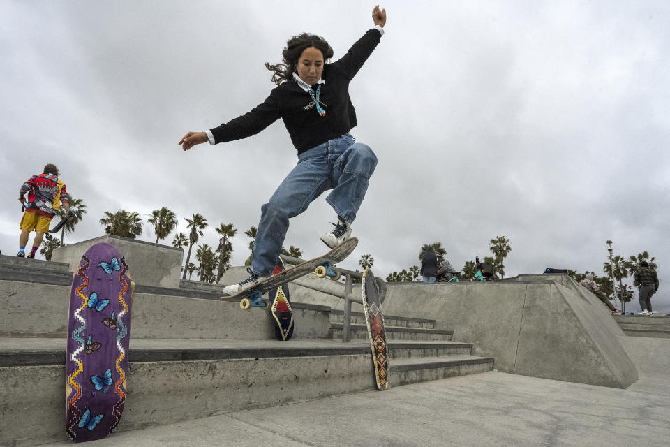 Expert skateboarder Di'Orr Greenwood, an artist born and raised in the Navajo Nation in Arizona and whose work is featured on the new U.S. stamps, rides her skateboard next to her artworks in the Venice Beach neighborhood in Los Angeles Monday, March 20, 2023. On Friday, March 24, the U.S. Postal Service is debuting the "Art of the Skateboard," four stamps that will be the first to pay tribute to skateboarding. The stamps underscore how prevalent skateboarding has become, especially in Indian Country, where the demand for designated skate spots has only grown in recent years. (AP Photo/Damian Dovarganes)