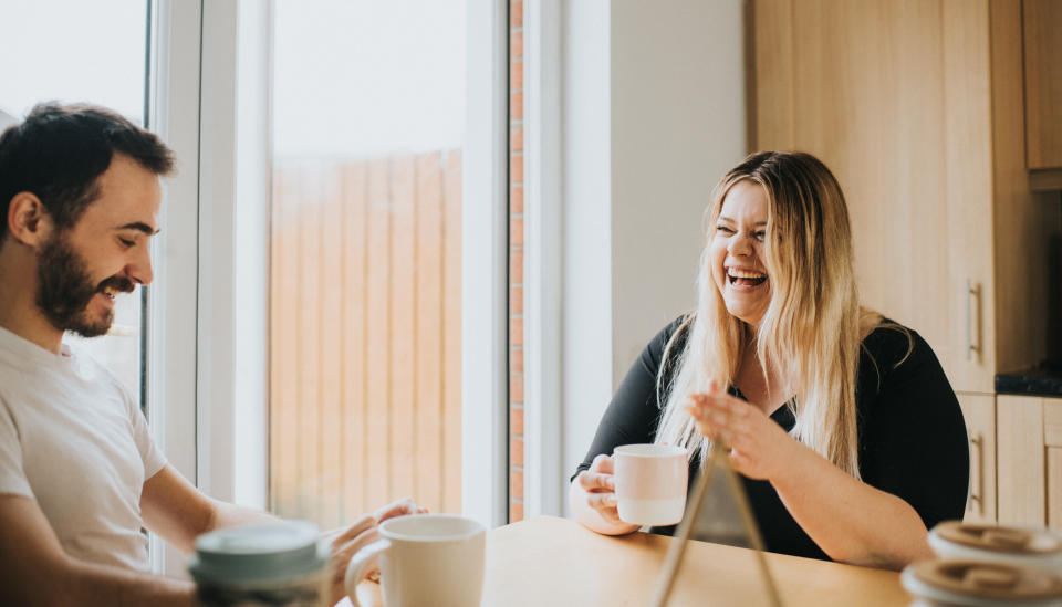 A couple sits at a dining table in the morning. They look happy and content in each other's company as they chat with eachother and laugh. They drink hot cups of tea/ coffee.