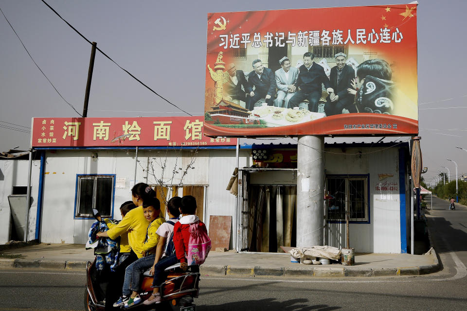 FILE - In this Sept. 20, 2018, file photo, an Uyghur woman uses an electric-powered scooter to fetch school children as they ride past a picture showing China's President Xi Jinping joining hands with a group of Uighur elders at the Unity New Village in Hotan, in western China's Xinjiang region. Australia and New Zealand on Tuesday, March 23, 2021 welcomed the United States, European Union, Canada and Britain taking joint action to impose sanctions on senior Chinese officials over human rights abuses in China’s far western Xinjiang region. (AP Photo/Andy Wong, File)