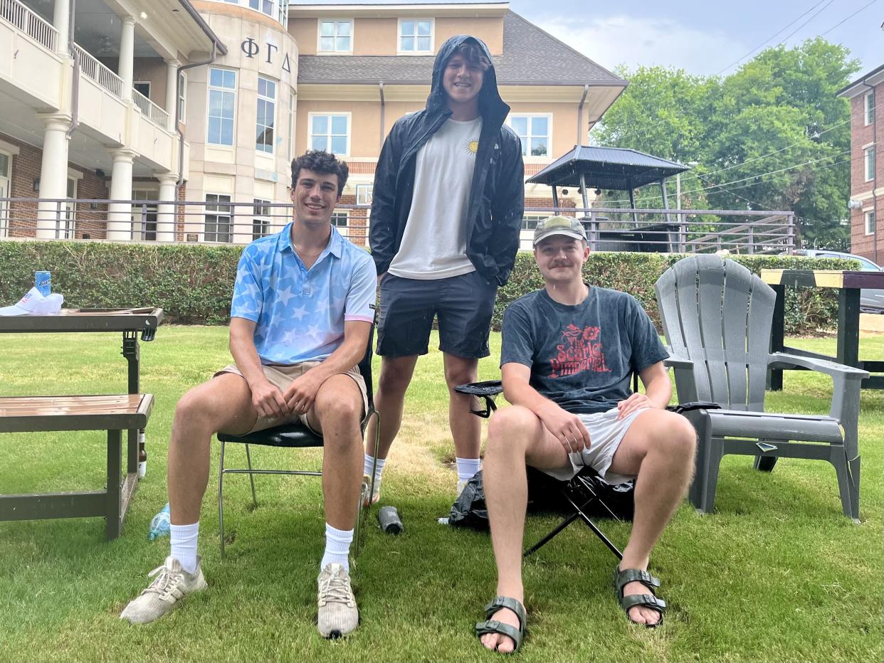 Three people smiling, two on lawn chairs, one standing behind them, on a lawn between buildings.