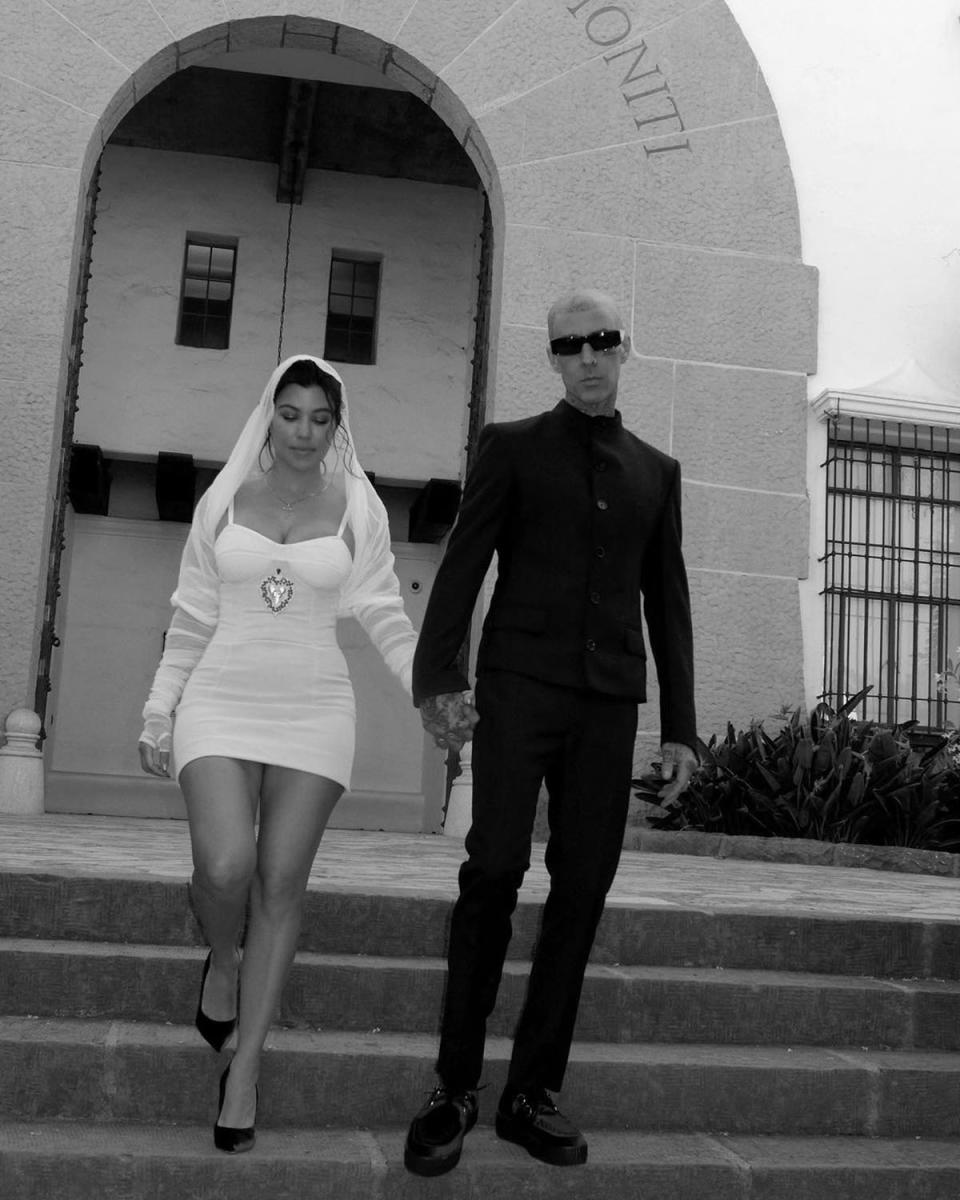Kourtney Kardashian in her bridal outfit, next to Travis Barker in a black suit and pants, holding hands while walking down some stairs.