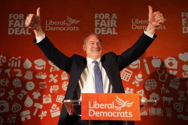 Sir Ed Davey, standing behind an orange Liberal  Democrat podium, gives a thumbs up gesture with both hands 