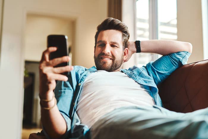 Man in a casual shirt lounging on a sofa, looking at his smartphone with a slight smile