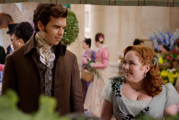 Nicola Coughlan and Luke Newton in period costume on the set of a historical drama, engaging in conversation amidst a crowd in similar attire