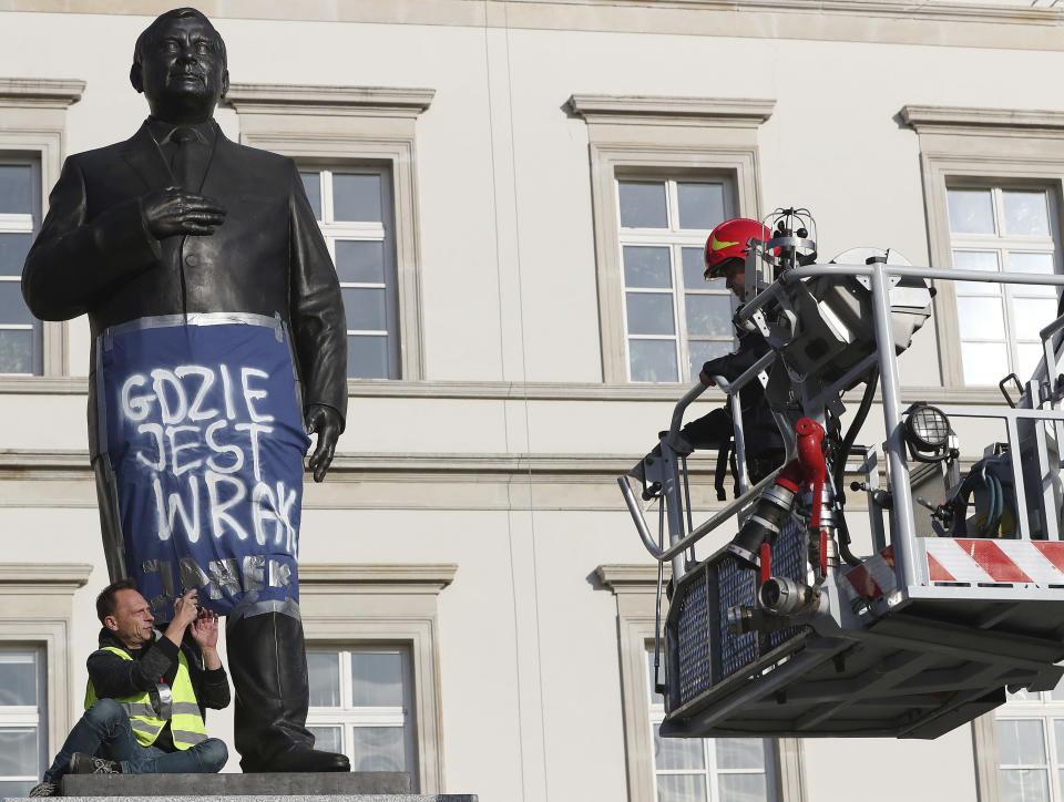 A man sits on a monument to Poland's late president Lech Kaczynski in downtown Warsaw, Poland, Friday, Oct. 11, 2019. He wrapped it in a banner reading 'Where is the Wreckage?' of the plane that crashed in 2010 in Russia, killing the president. Just two days ahead of parliamentary elections, this is criticism of the ruling Law and Justice party, led by the late president's twin Jaroslaw Kaczynski, which has vowed to bring the wreckage back from Russia, where it remains. (AP Photo/Czarek Sokolowski)
