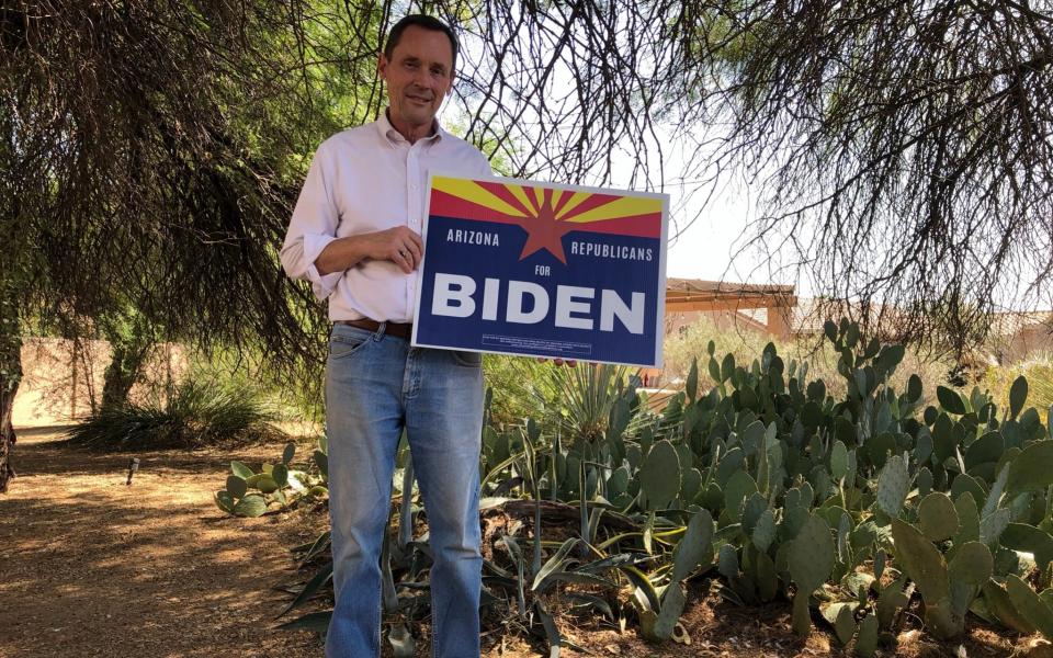A tall, white, older man with close-cropped black hair, jeans and wearing a white buttoned shirt with its sleeves rolled up, holds a sign that reads "ARIZONA REPUBLICANS FOR BIDEN". He is standing amid cacti in a bright back yard, with the low shape of his house just visible through bushes and trees behind him - Laurence Dodds/Telegraph