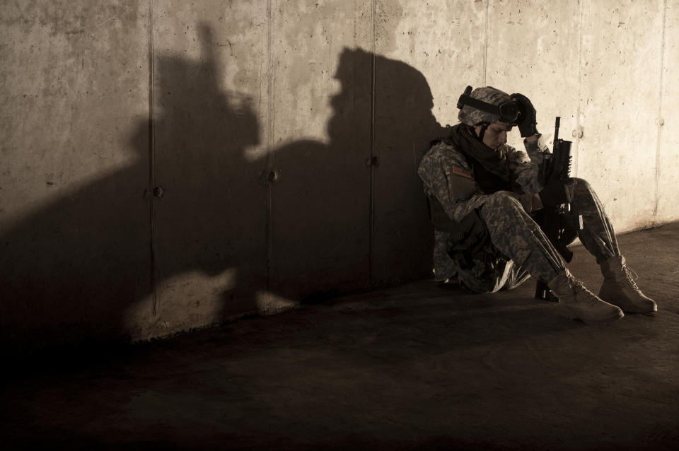 A soldier in military gear sits on the ground, head down, holding a rifle. The shadow on the wall shows the outline of Batman