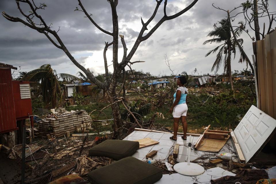 A woman stands on a tiled floor of what appears to have been a house, surrounded by fallen palm trees, destroyed structures and endless debris.