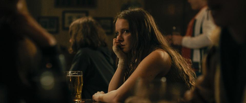 A woman leans her chin on her hand as she sits in a bar with a glass of beer.