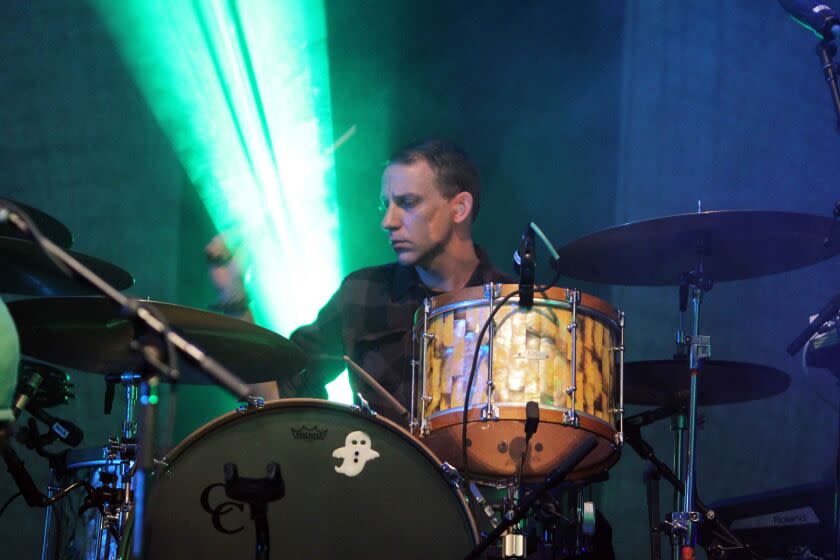 A man with short hair playing drums on a stage illuminated with green light
