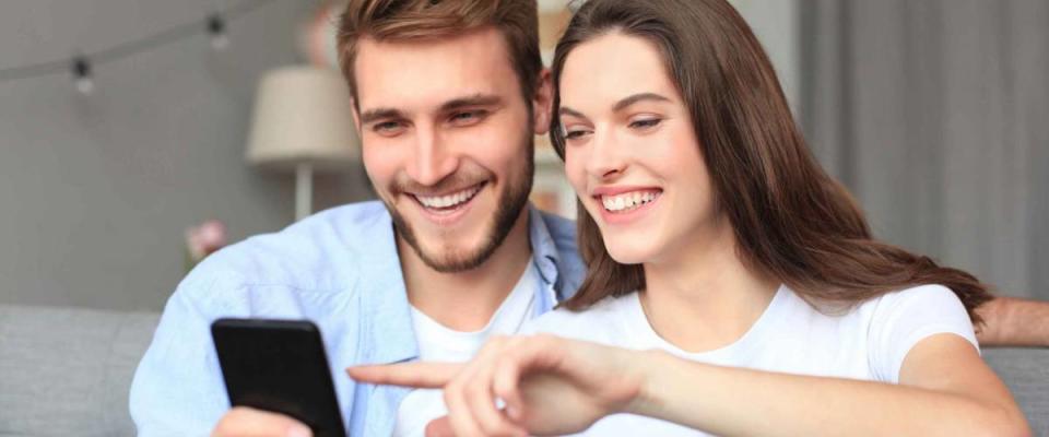 Young couple watching online content in a smart phone sitting on a sofa at home in the living room.