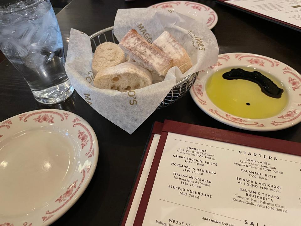 Dark wooden table with a Maggiano's menu, a basket of sliced bread, and a plate of olive oil