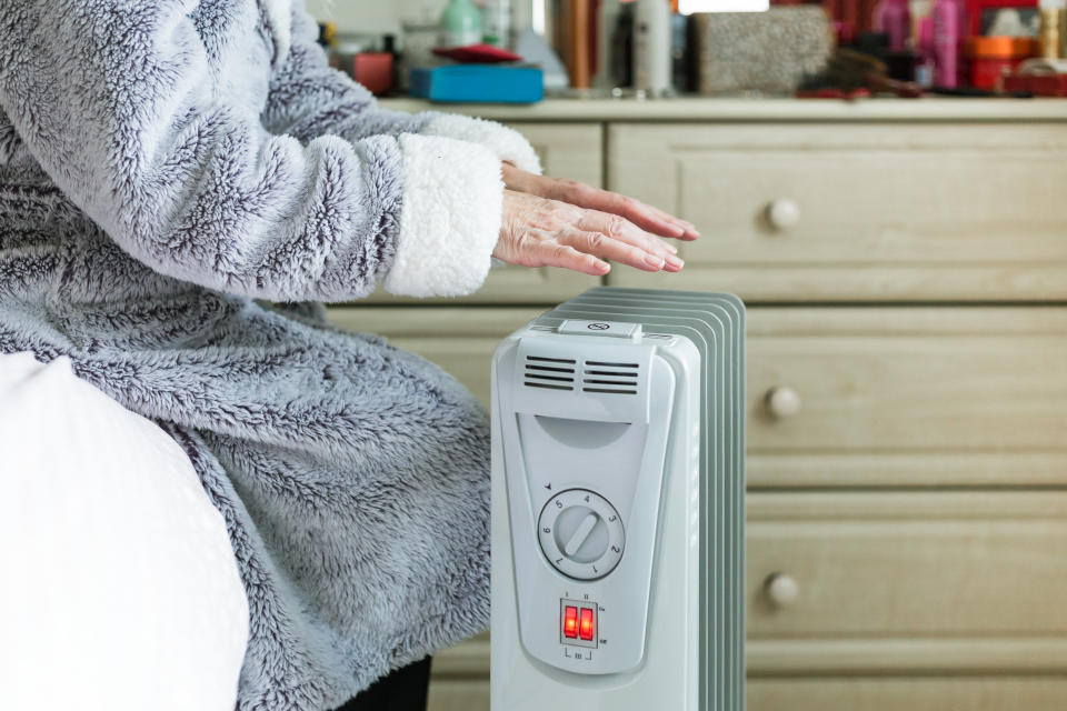 A retired senior woman in her 70s sits at home inside her cold house in winter. It is so cold that she is wrapped up in warm winter clothing, and is holding her hands over an electric heater for some extra warmth and comfort. Selective focus with room for copy space.