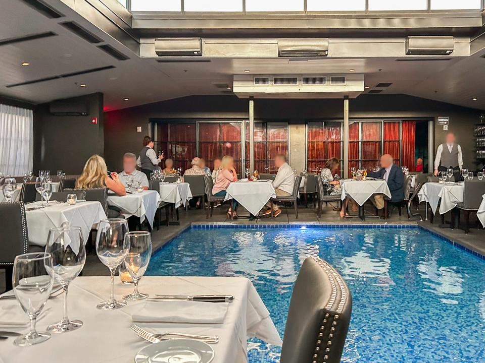 People dine at a restaurant with white table cloths, gray, leather chairs, and a rectangular pool diving the dining room