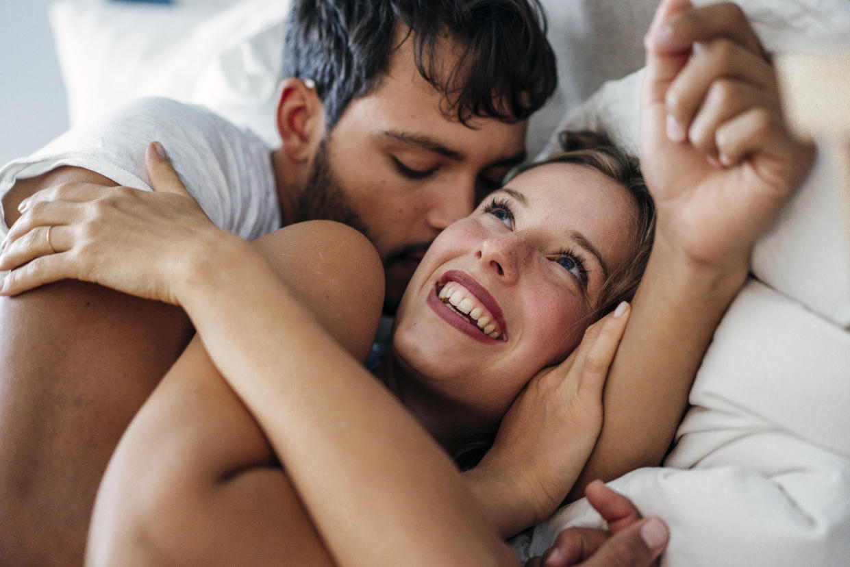 A young couple spoon in bed, a man gives his partner a kiss on her cheek as she smiles up at him