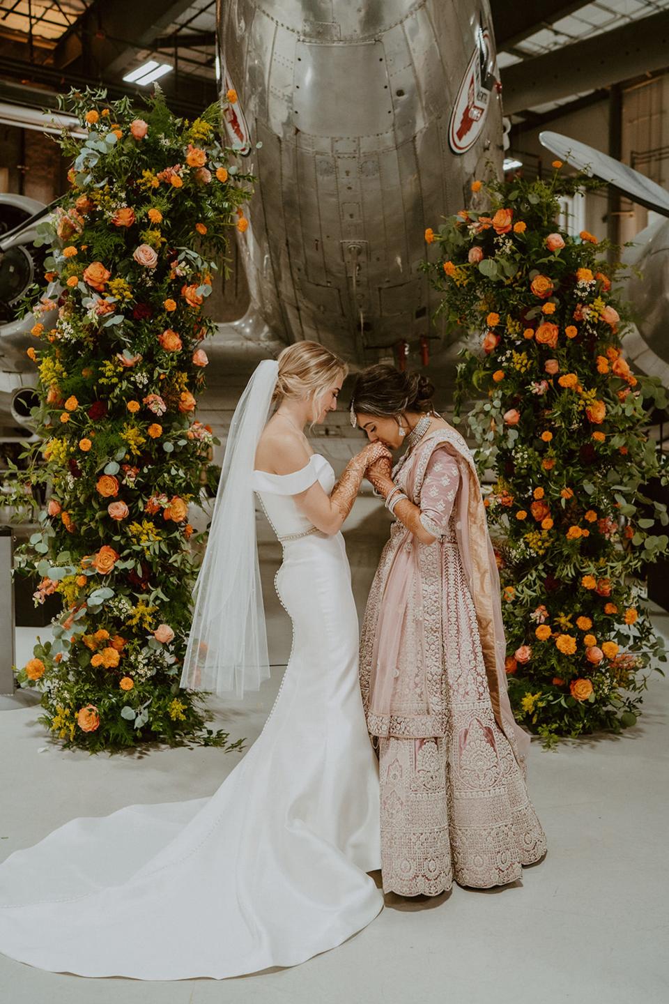 A bride holds her wife's hands and kisses them in front of a floral arch and airplane.
