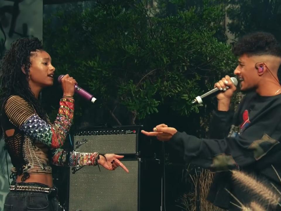 Willow Smith (left) and Tyler Cole (right) facing each other as they sing into handheld mics. Willow has a half-up half-down do, and is wearing a slightly cropped and fitted long sleeve tee with mismatched patterns. Tyler Cole is wearing a baggier long sleeve tee. They appear to be singing to each other.