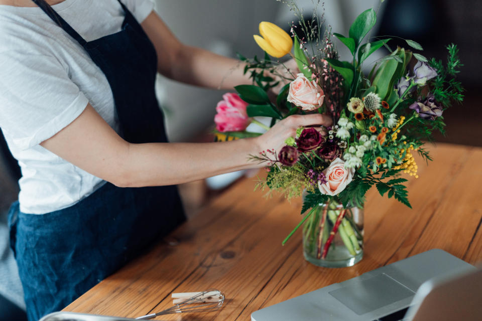 A person arranges a bouquet of flowers on a wooden table next to a laptop. The image relates to work and money, suggesting a job or business in flower arranging