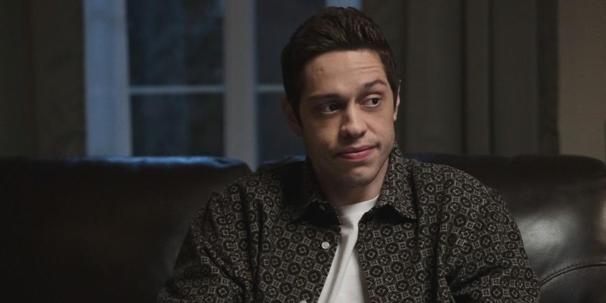 pete davidson wearing a black and white buttoned shirt and a white t shirt, sitting on a couch