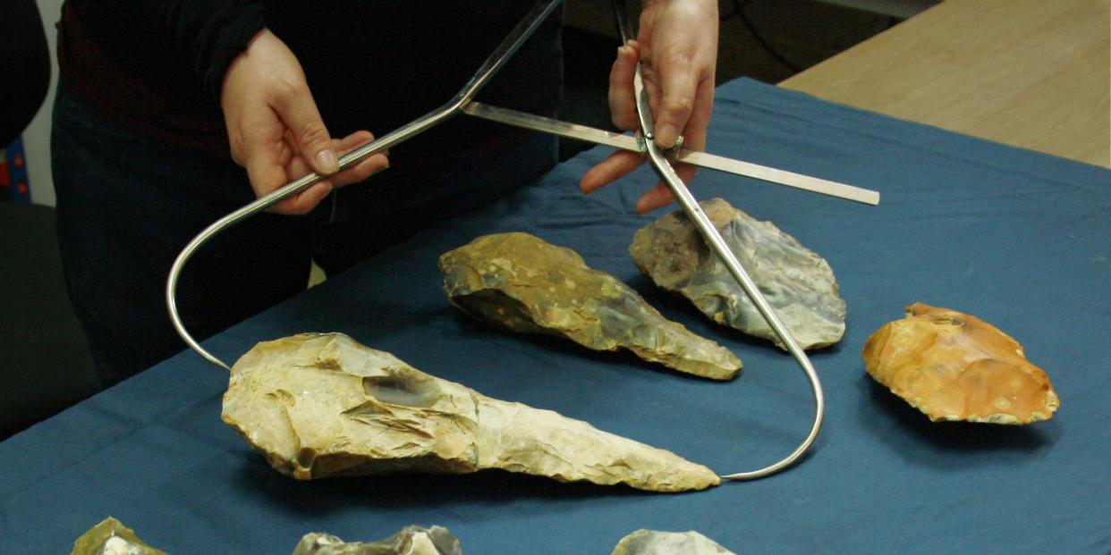 Ingrey uses a tool to measure the length of the large hand axe found at the English archeological site. Other artifacts are laid out on the table near the large handaxe.