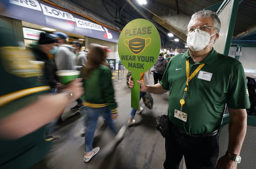 John Abercrombie, an usher for the Oakland Athletics, holds up a sign advising people to wear masks, prior to a baseball game between the Ahletics and the Houston Astros on Friday, April 2, 2021, in Oakland, Calif. (AP Photo/Tony Avelar)