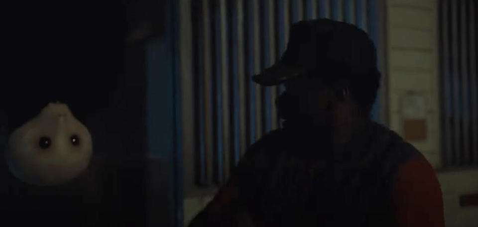 A man wearing a cap sits inside a dimly lit room, with a white, ghostly figure with big, dark eyes floating beside him