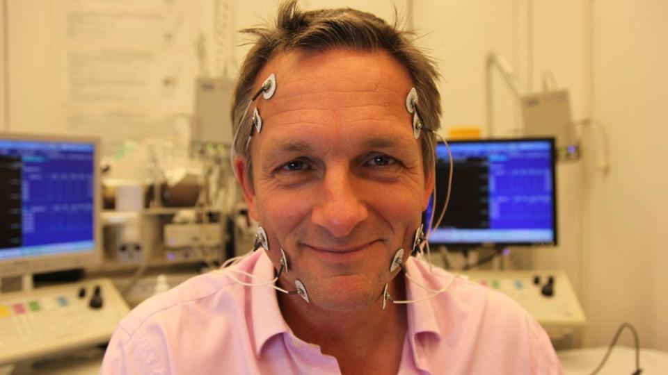 Michael Mosley in a promotional photo for a past TV show, smiling with medical sensors attached to his face and computer monitors behind