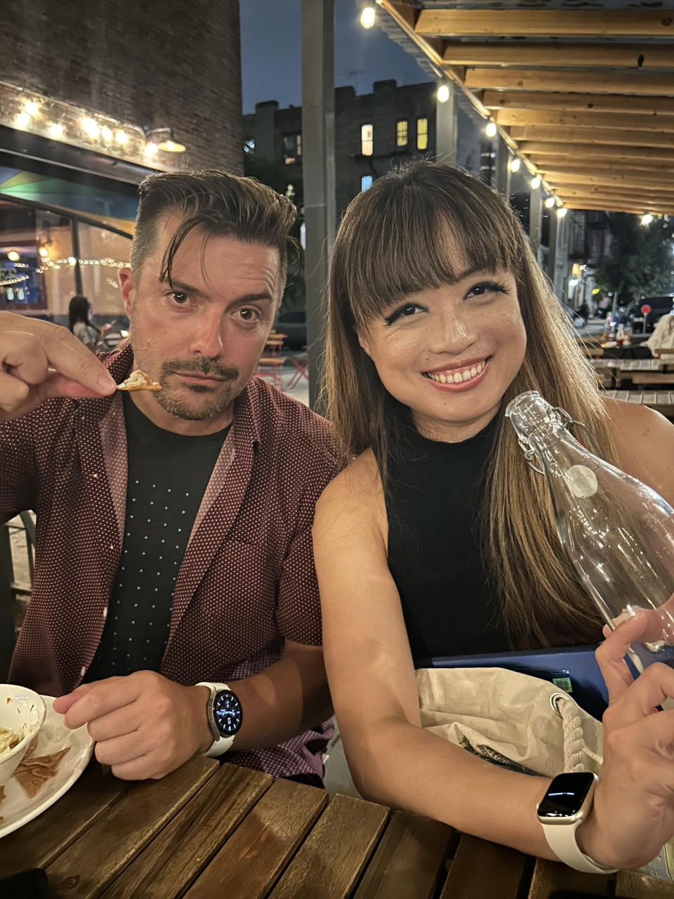 <p>A photo from the iPhone 14 showing a man and woman posing with food and a water tumbler at an outdoor restaurant at night.</p>

