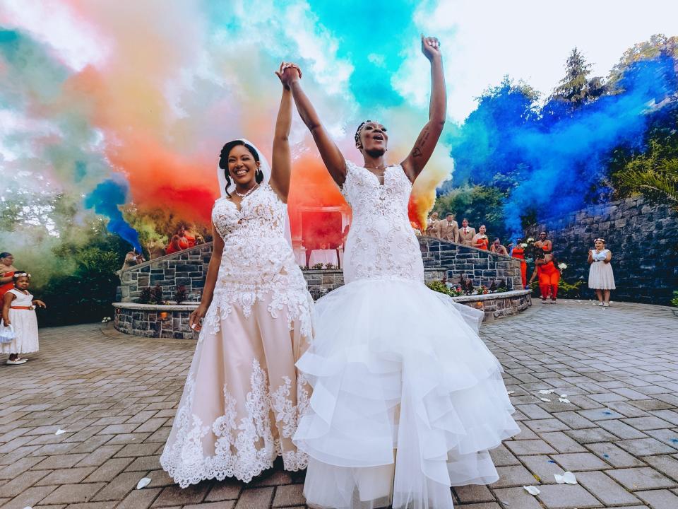 Two brides hold hands as rainbow-colored smoke explodes behind them.
