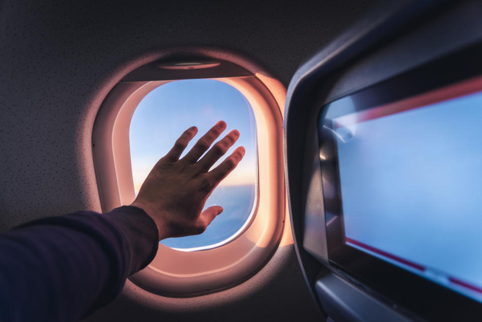 Personal perspective of human hand touching the airplane's window while flying above the sea at sunrise