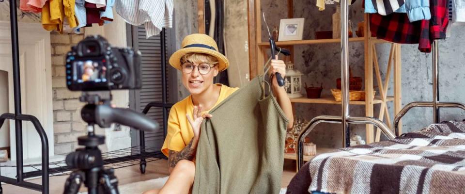 Young woman fashion blogger influencer wearing hat and eyeglasses sitting on fluffy carpet at urban stylish apartment recording video vlog on digital camera telling clothing tips positive