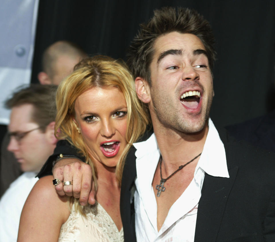 Britney and Colin posing playfully on a red carpet; she's in a sleeveless outfit and he's in a suit with an open shirt and his arm is around her shoulders