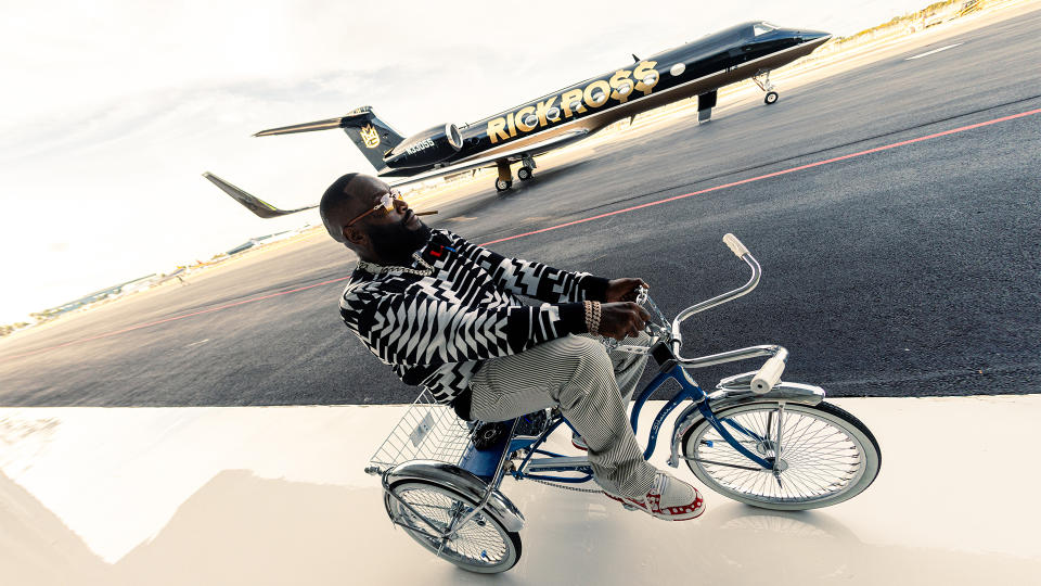 Rick Ross wears his own Louis Vuitton sweater and sneakers, Exclusive Games pants, Vobara chain and bracelets, and Cartier glasses while riding his Schwinn retro tricycle.