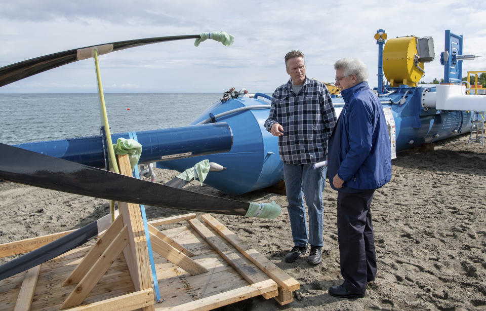 In this Tuesday, July 16, 2019, photo released by the Alaska Governor's office, Gov. Mike Dunleavy, left, talks with ORPC CEO, Co-Founder, and Chairman Chris Sauer in front of a Riv-Gen Power System turbine on the bank of the Kvichak River in Igiugig, Alaska. A tiny Alaska Native village is adopting an emerging technology to transform the power of a local river into a renewable energy source. (Austin McDaniel/Alaska Governor's Office via AP)