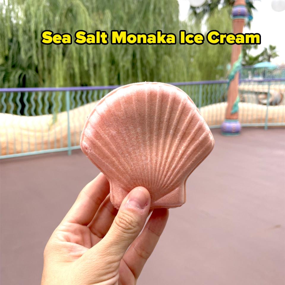 A hand holds a pink seashell-shaped object in an outdoor setting with greenery and themed lamp posts in the background