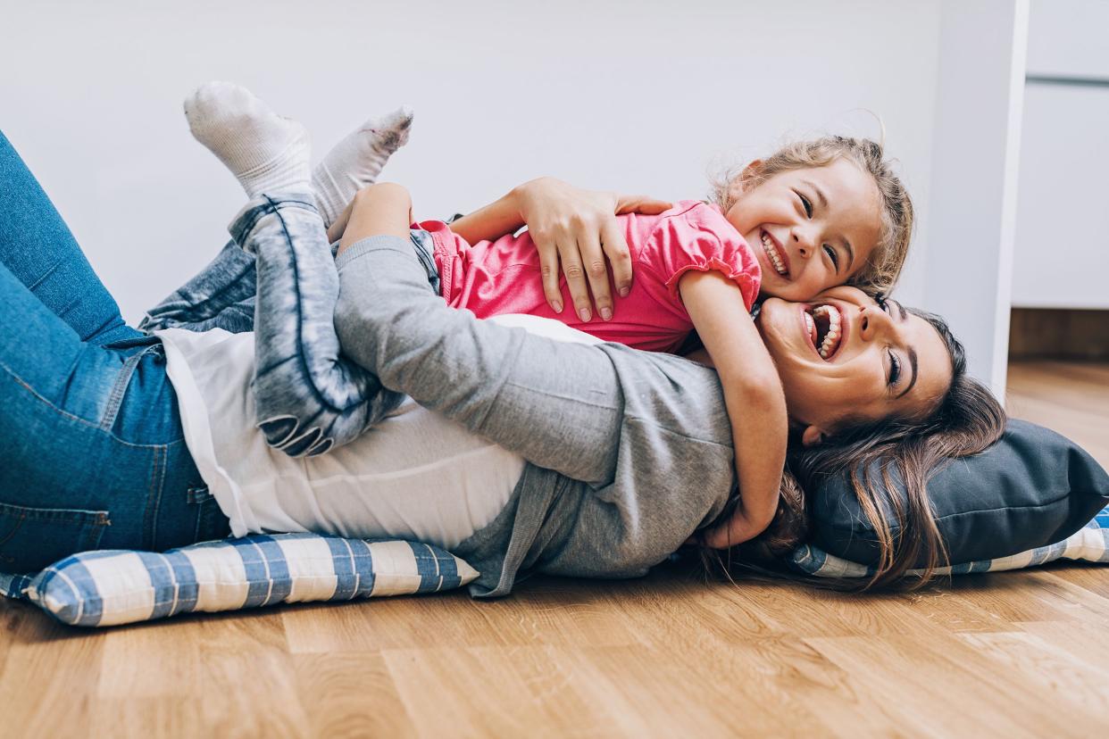Laughing young woman laying down hugging a smiling little girl on pillows on the wooden floor, white wall in the background