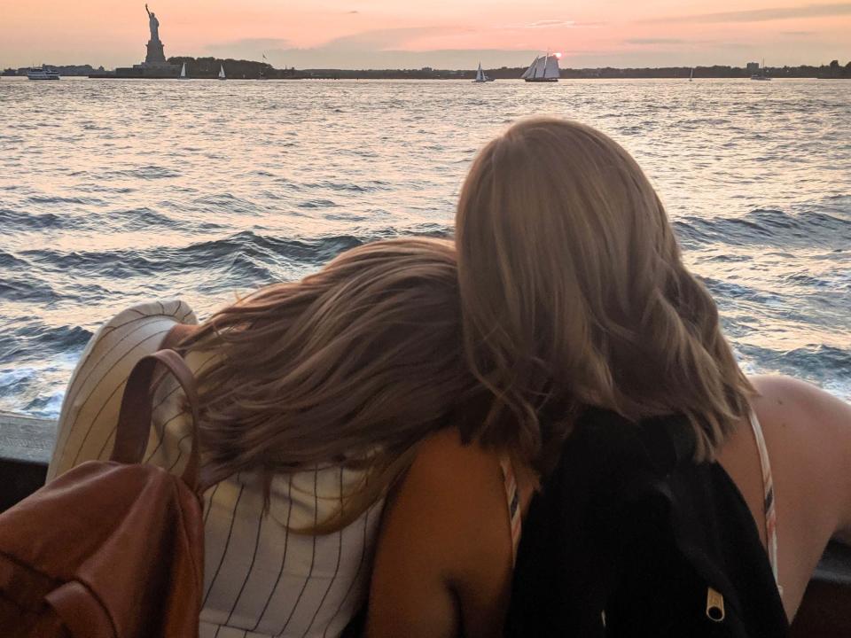 Two people face away from the camera with the sea and the Statue of Liberty in front of them during sunset
