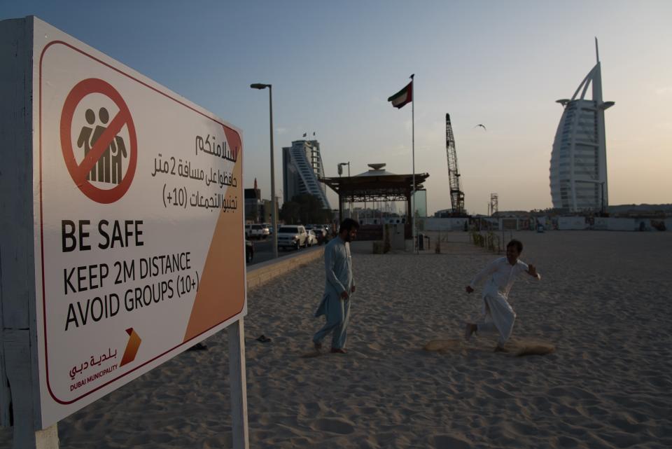 Two laborers play tag near a sign warning people to maintain a distance from each other over the outbreak of the new coronavirus in front of the sail-shaped Burj Al Arab luxury hotel in Dubai, United Arab Emirates, Friday, March 20, 2020. The United Arab Emirates has closed its borders to foreigners, including those with residency visas, over the coronavirus outbreak, but has yet to shut down public beaches and other locations over the virus. (AP Photo/Jon Gambrell)