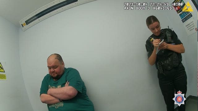 Gavin Plumb in a green T-shirt sits with his arms crossed in a police interview room while a female officer stands behind him making notes