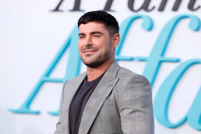 Zac Efron stands smiling at an event, wearing a checkered blazer over a dark shirt, with a "A Family Affair" sign in the background