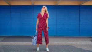 Pretty blond woman wearing burgundy jumpsuit and sunglasses while walking against the blue wall and holding cerulean tote bag