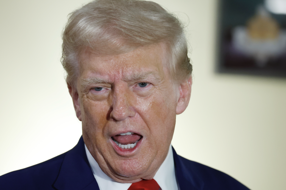 Donald Trump speaking, wearing a dark suit and red tie, with an expressive face