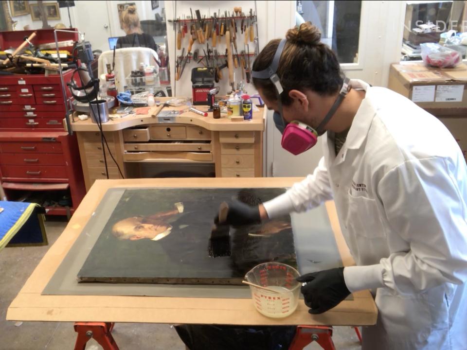 An conservator holding a brush puts varnish onto a painting.