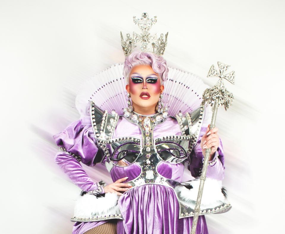 Drag queen in elaborate costume, including a jeweled crown, ruffled collar, ornate silver detailing, and a long velvet gown with fishnet stockings, posing with a scepter