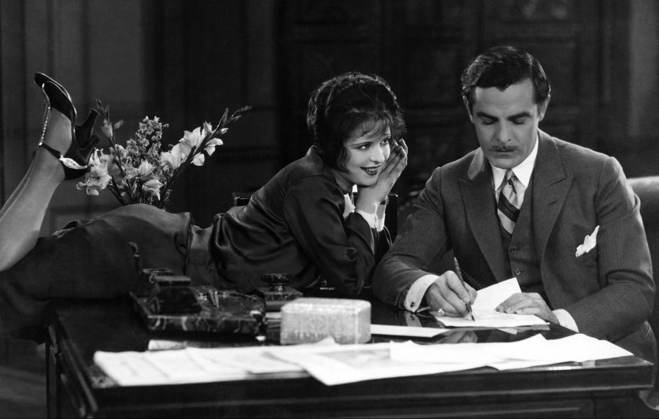 clara bow and antonio moreno in a scene from the movie it, she lies on her stomach with her legs up in the air and smiles at him, he sits at a table writing, several other papers are on the table as well