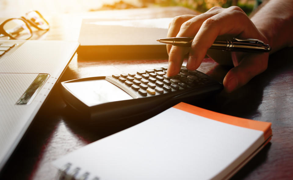 Hand holding pen and typing on calculator that's on a desk next to a steno pad and a computer.