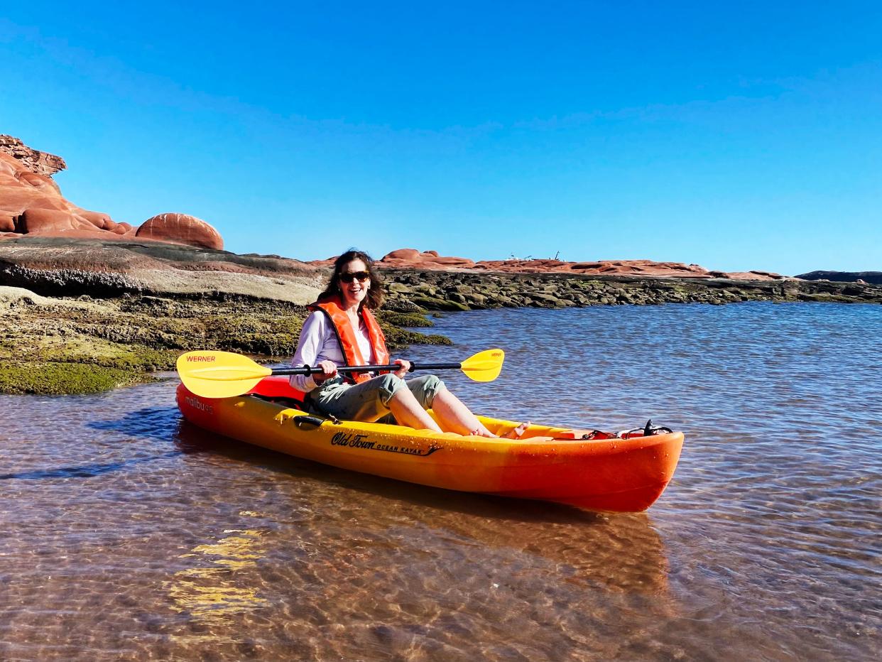 A woman in an orange kayak holding a yellow paddle wades in shallow water.