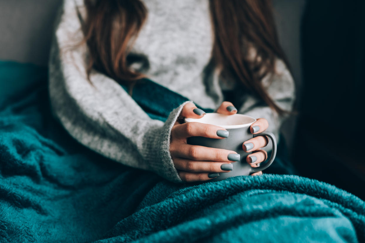 Frau sitzt in Pullover und Decke und hält eine Tasse Tee in den Händen